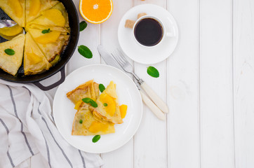 Crepe suzette with orange sauce and a cup of coffee on a white wooden background. Tasty and fragrant breakfast.