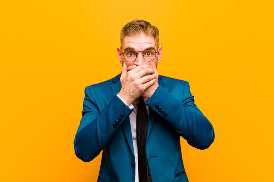 young red head businessman covering mouth with hands with a shocked, surprised expression, keeping a secret or saying oops against orange background