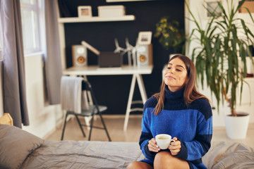 Charming brunette in blue sweater sitting in bedroom in morning and drinking coffee.