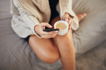 Top view of caucasian woman using smart phone and holding cup of coffee while sitting on bed in morning.