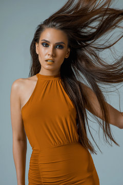 Young Beautiful Brunette With Blue Smokey Eyes And Yellow Lips, Professional Makeup, Contouring, A Female In An Orange Dress Throws Long Black Hair Up On A Blue Background