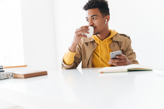 Image of african american guy holding smartphone and cup in office