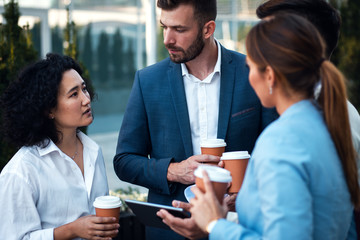 Group of coworkers having a coffee brake together, standing outside in front of office buildings and talking.