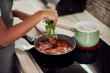 Mixed race woman in apron standing next to stove, stirring tomato sauce and adding rocket. On stove is pot with boiling pasta.