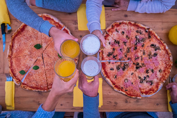 close up of hands with glass with beer and orange juice clinking together at the centre of the table - two big pizzas on the wooden table