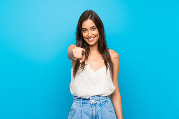 Young woman over isolated blue background points finger at you while smiling