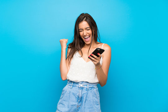 Young woman over isolated blue background with phone in victory position