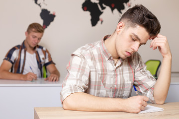 Students in classroom with world map on the wall on background. Young guy writes an abstract or does a test. Education, university, learning, coworking concept