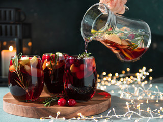 Female hand pours winter sangria in glasses with fruit slice, cranberry and rosemary. Dark christmas holiday background with candle, decoration lighting chain. - obrazy, fototapety, plakaty