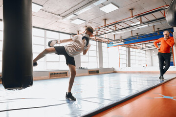 young boxer learning to blow with leg, serious coach standing in the background of the photo. checking the boy's workout,