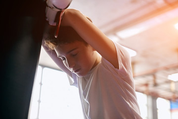 boy is being tired after workout. kid having a rest after training , leaning on the heavy punch bag in boxing gym. close up side view photo. relaxation, tiredness, failure