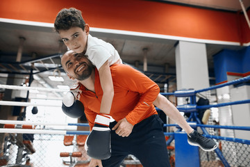 cheerful young bearded man giving piggyback ride to smiling kid in boxing gloves at gym, free time, spare time, lifestyle