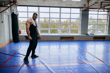 tired muscular bodybuilder with water bottle and towel on his shoulder looking down having a rest. copy space. full length photo. tiredness, exhaustion, punching bag in the background of the photo © the faces
