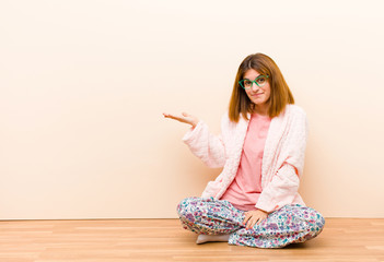 young woman wearing pajamas sitting at home feeling happy and smiling casually, looking to an...