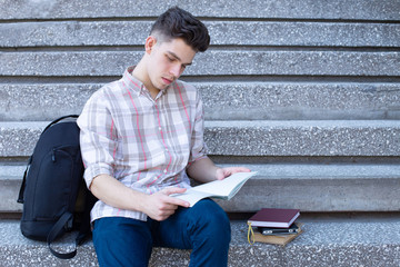 Young guy student sitting on the stairs of a school, college or university and reading a precis