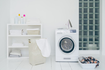 Laundry room with washing machine, dirty clothes in basket, iron and little shelf with neatly folded linen. Domestic room interior. Washing concept