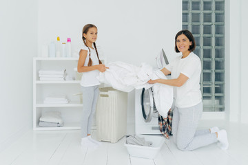 Glad housewife does washing with little adorable helper. Mother and daughter wash clothes in laundry room, load linen in washer. Woman stands on knees near washing machine. Housework concept