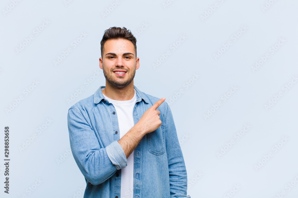 Wall mural young hispanic man smiling cheerfully, feeling happy and pointing to the side and upwards, showing object in copy space against blue wall