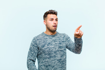 young hispanic man feeling shocked and surprised, pointing and looking upwards in awe with amazed,...