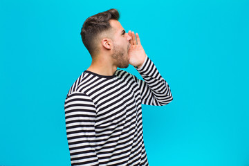young hispanic man yelling loudly and angrily to copy space on the side, with hand next to mouth...