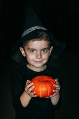 Portrait of a little boy dressed in black clothes and a black magic hat, holds a small orange halloween pumpkin on a black background. Halloween 2019