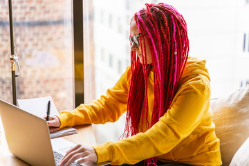 Girl with pink long hair, dreadlocks works at computer in cafe. The concept of remote work,...