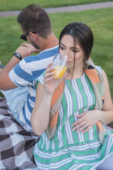 Sexy brunette pregnant woman in park, lay on her husband and drinking juice. Summer day, sunset