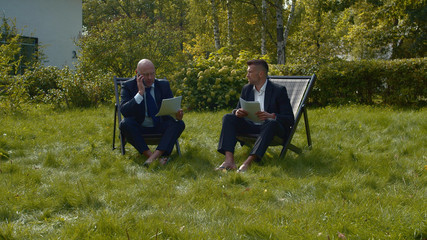 Men in suits sit in courtyard with papers and talk.