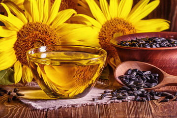 Rural still-life - the sunflower oil in glass bowl with flowers of sunflower (Helianthus annuus) and raw seeds against the background of an wooden wall, closeup