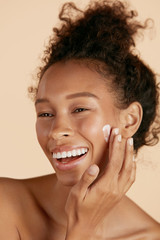 Face skin care. Woman applying cosmetic cream on clean hydrated skin portrait. Beautiful happy smiling african american girl model with natural makeup applying facial moisturizer, beauty product