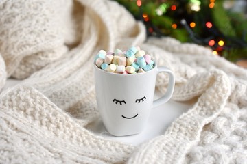 Fototapeta na wymiar Warm, cozy holiday season composition, cup with smiley face, cacao and mini marshmallows, Christmas tree with lights on background.