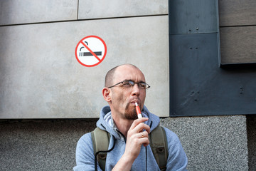 Portrait of caucasian man smoke electronic cigarette Iqos under sign No smoking