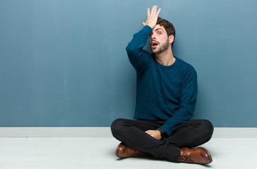 young handsome man sitting on floor raising palm to forehead thinking oops, after making a stupid...
