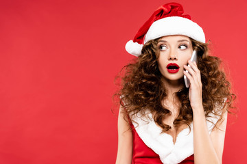 attractive shocked woman in santa costume talking on smartphone, isolated on red