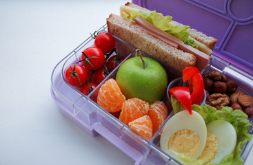 Close up of lilac lunch box with useful food for lunch and snack: sandwich, vegetables, fruits, nuts and eggs. Concept of healthy food, snack for adults and kids. White background, copy space