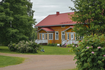 Fototapeta na wymiar Finnish countryside with yellow wooden finnish house at strawberry farm and green bush with pink flowers at foreground, summer day