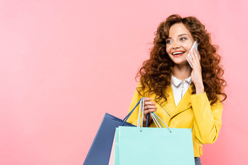 Fototapeta premium beautiful curly girl holding shopping bags while talking on smartphone, isolated on pink