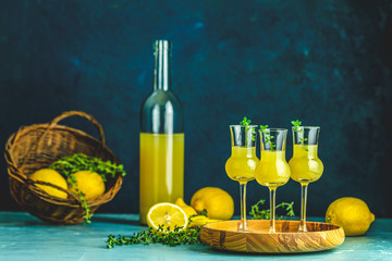 Limoncello with thyme in three grappas wineglass in wooden tray, fresh lemon in basket on dark blue...