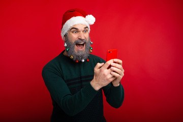 Amazed man with decorated beard and wearing santa claus hat and looking shocked at smartphone