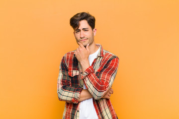 young handsome man looking serious, thoughtful and distrustful, with one arm crossed and hand on...