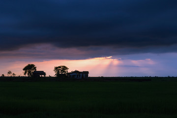 Naklejka na ściany i meble the last rays of the sun break through the dark evening clouds and fall on a cozy house in the middle of a huge field