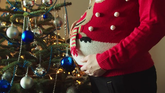 Young 9 months pregnant mother cuddling her tummy in the Christmas sweater in front of the decorated Christmas three