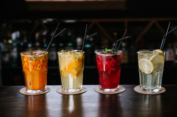 Different types of summer drinks in glasses, cubes of ice and slice of fruits on dark wiooden table.