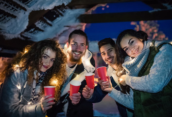 Group of young friends outdoors in snow in winter at night, holding drinks.