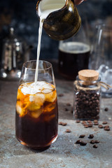 Ice coffee cold summer drink in a tall glass with pouring cream and coffee beans on a stone background.