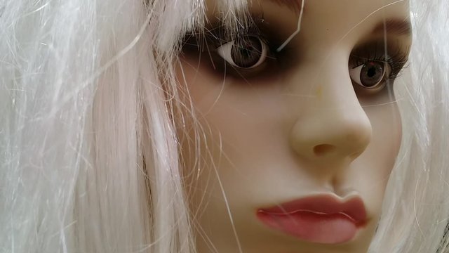 Movement in footage of closeup female mannequin face with dark makeup around her big beautiful eyes, big red lips, super smooth skin, and fake shiny white blond hair moving in the wind.