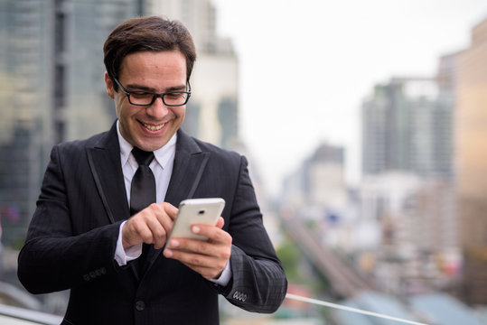 Happy handsome Persian businessman using phone in the city
