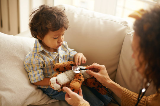 Portrait Of Cute Mixed Race Male Child Sitting On Sofa With Plush Toy Playing With Unrecognizable Mother Pretending To Be Doctor Using Stethoscope As If Listening To Internal Sounds Of Stuffed Tiger