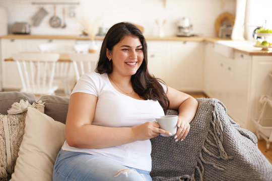 Portrait of cheerful charismatic young overweight female with big breast and long black hair posing in stylish kitchen interior, having coffee on comfortable couch and laughing at funny joke