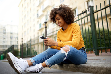 cool young african american woman sitting outside on street with cellphone - Powered by Adobe